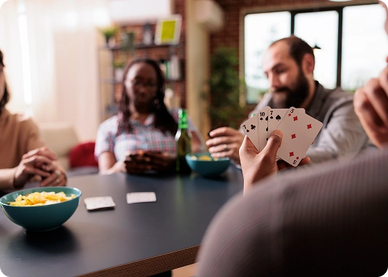 Vier mensen zitten rond een tafel te kaarten in een gezellige kamer. De focus ligt op een hand met kaarten. Er staan schalen met chips en er heerst een ontspannen sfeer.