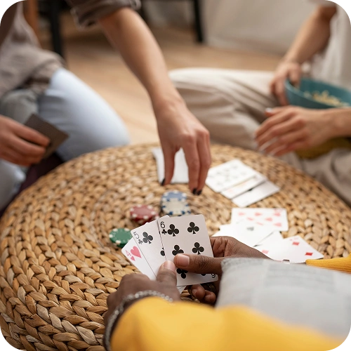 Mensen zitten rond een gevlochten tafel te kaarten. De ene persoon houdt de speelkaarten vast, terwijl de andere naar de pokerfiches reikt. De sfeer is ontspannen en gezellig.
