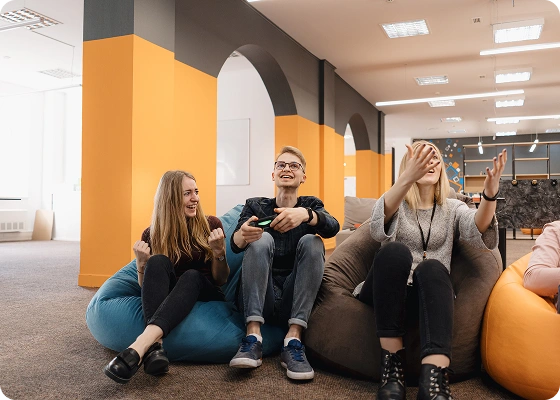Drie jongvolwassenen zitten op zitzakken in een modern, helder verlicht kantoor met oranje accenten. Ze glimlachen en lijken ontspannen te kletsen.