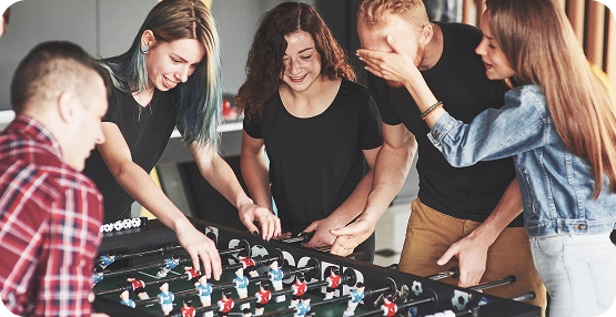 Een groepje van vijf vrienden speelt binnen tafelvoetbal; ze lachen en zijn helemaal in hun spel verdiept. De sfeer is levendig en vrolijk, met informele kleding en natuurlijk licht.