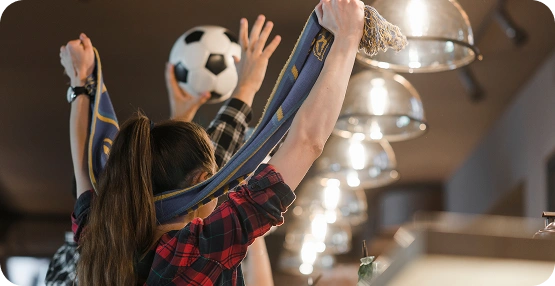 Twee mensen in een bar vieren iets terwijl ze een sjaal en een voetbal vasthouden. De sfeer is levendig, met warme verlichting en een gevoel van opwinding.